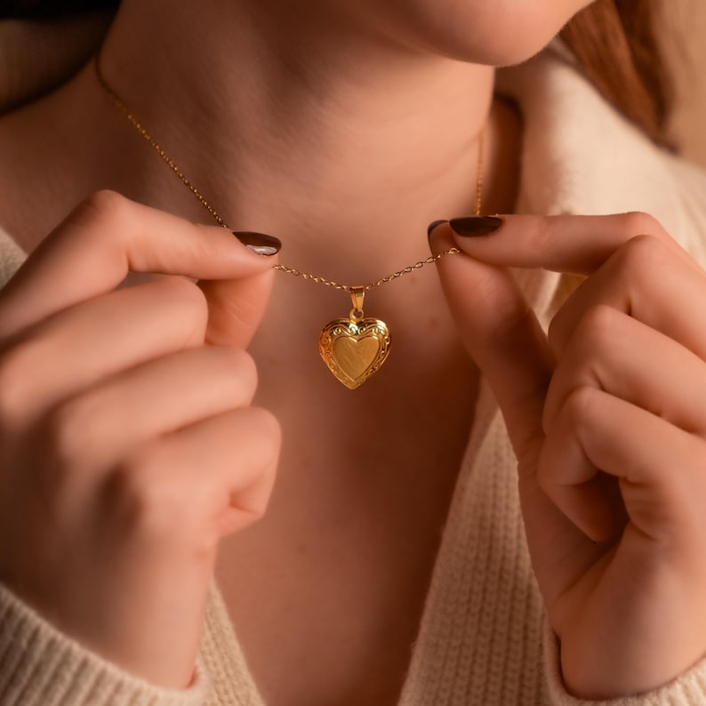 May include: A gold heart-shaped locket necklace with a delicate chain. The locket has a detailed design and is worn by a person.