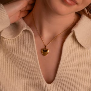 May include: A gold heart-shaped locket necklace on a person wearing a cream-colored sweater.