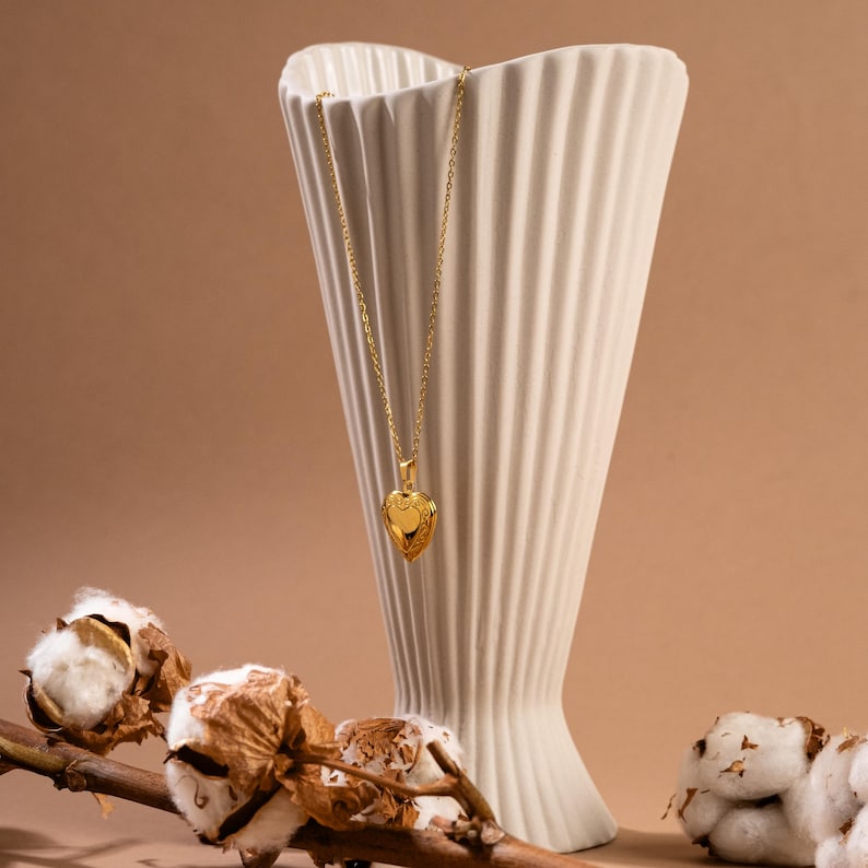 May include: A gold heart-shaped locket necklace hanging from a white ceramic vase with a textured surface. The necklace is on a chain and the locket has a detailed design. The vase is on a brown surface with a cotton branch.