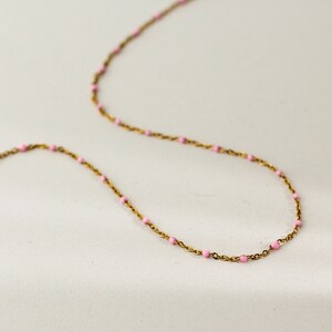 May include: A delicate gold-toned necklace with small, evenly spaced pink beads. The chain is thin and the beads are a soft pastel pink, creating a minimalist and elegant aesthetic. The necklace is displayed on a light background.
