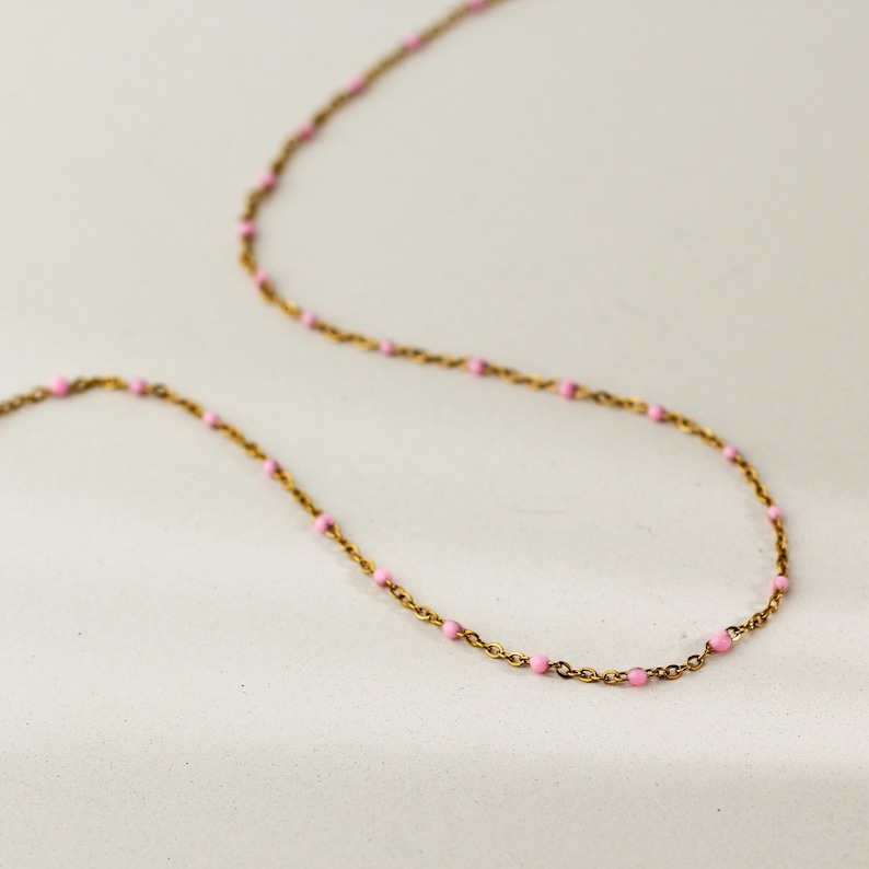 May include: A delicate gold-toned necklace with small, evenly spaced pink beads. The chain is thin and the beads are a soft pastel pink, creating a minimalist and elegant aesthetic. The necklace is displayed on a light background.