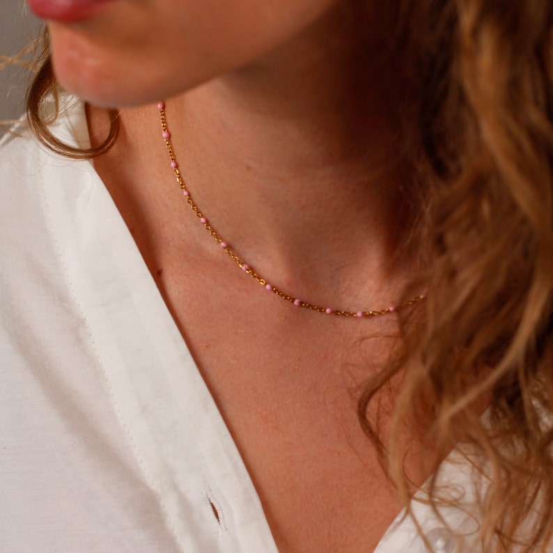May include: A delicate gold necklace with small, evenly spaced pink beads. The necklace is worn against a white shirt, creating a contrast that highlights the jewelry. The chain is fine and elegant.