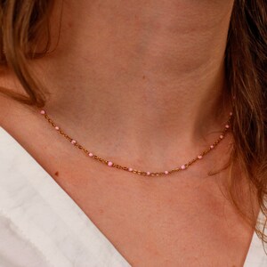 May include: A delicate gold-colored necklace with small, evenly spaced pink beads. The necklace is worn around the neck, and the background is a white shirt and skin.
