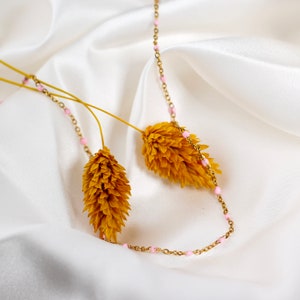 May include: A delicate gold-toned necklace with small pink beads is draped over a white satin surface. The necklace is accented by dried yellow flowers, creating a soft, elegant aesthetic. The chain is fine and the beads are evenly spaced.