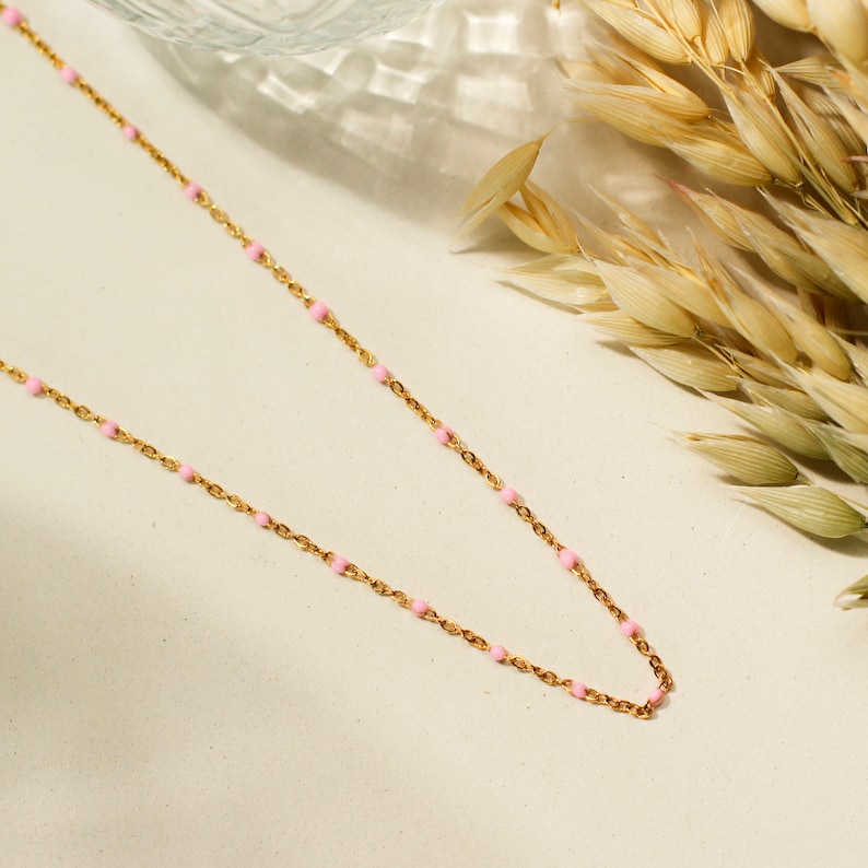 May include: A delicate gold-toned necklace featuring small, evenly spaced pink beads. The necklace is displayed on a light-colored surface, with a sprig of dried wheat in the background. The chain is fine and elegant.