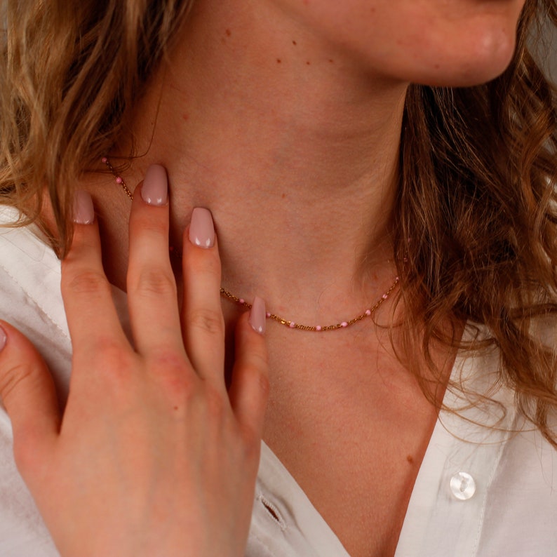 May include: A delicate gold necklace with small pink beads. The necklace is worn around the neck and is visible against a white shirt. The hand is touching the necklace.