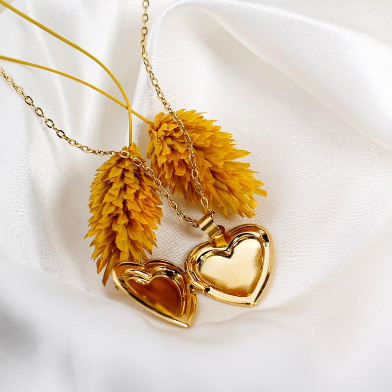 May include: Two gold heart-shaped locket necklaces with delicate chains. The lockets are open and the chains are draped over dried yellow flowers.