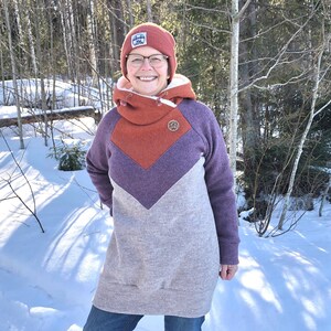 May include: A woman wearing a purple, orange, and gray hooded sweatshirt with a brown button. She is wearing blue jeans and a red beanie with a white logo. She is standing in a snowy forest.