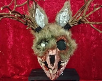 May include: A handmade, eerie mask with a rabbit-like face, featuring one white eye and a dark eye socket. The mask is adorned with faux fur, wooden branches, and a skeletal jaw. The mask is set against a red backdrop.