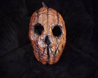 Puede incluir: Una máscara de calabaza marrón y negra con una cara tallada. La máscara tiene dos agujeros para los ojos y una boca tallada. La máscara está hecha de papel maché y tiene una superficie texturizada.