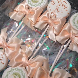 Puede incluir: Cake pops decorados con lazos de satén color melocotón. Algunos tienen chispas naranjas, otros presentan un diseño verde y blanco con el texto "Eid Mubarak". Presentados en una caja transparente.