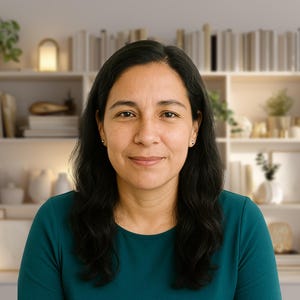 May include: A woman with dark hair and a teal top smiles at the camera. Behind her is a white bookshelf filled with books, plants, and decorative items. The background is softly lit, creating a warm and inviting atmosphere.
