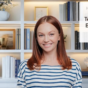 May include: A woman with long red hair smiles at the camera. She is wearing a blue and white striped shirt. The background is a white bookshelf with books and decorative items.