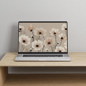 May include: A laptop displays a floral wallpaper featuring cream-colored flowers with dark centers against a beige background. The laptop is on a light brown wooden table, set against a neutral wall.