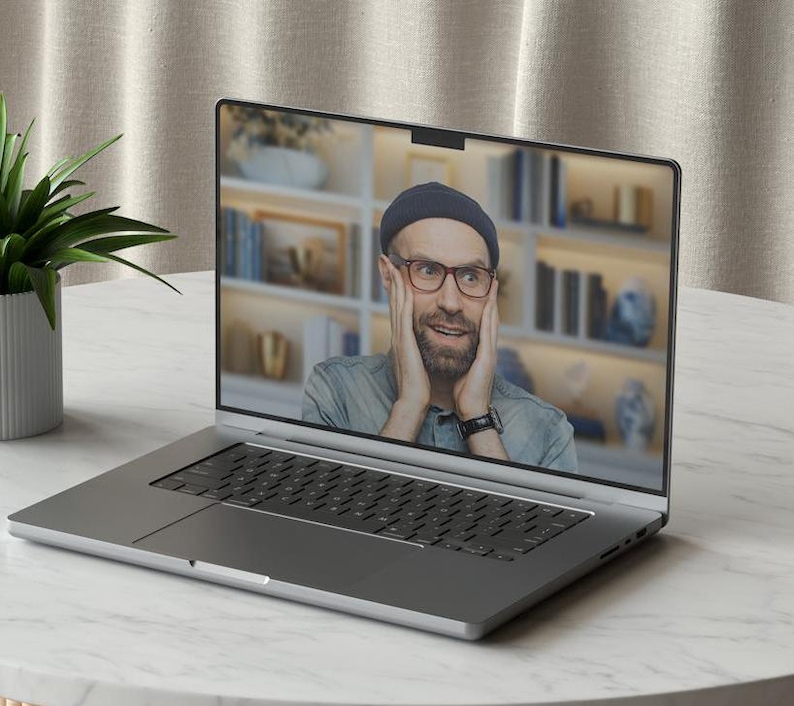 May include: A silver laptop computer with a man on the screen looking surprised with his hands on his cheeks. The man is wearing a blue denim shirt, a black beanie, and glasses.