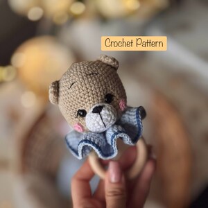 May include: Crocheted teddy bear rattle with a blue and white ruffled collar, attached to a wooden ring. Crochet Pattern.