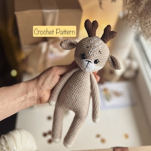 Puede incluir: Un juguete de crochet de ciervo beige con cuernos marrones. El ciervo tiene ojos blancos y una nariz blanca. El juguete está siendo sostenido por una mano.