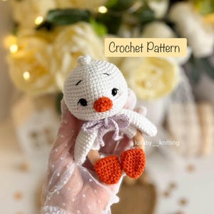 May include: A handmade white crochet duckling with an orange beak and feet, and a lavender bow. The duckling has black eyes and is holding a crochet pattern. The text "Crochet Pattern" is visible.