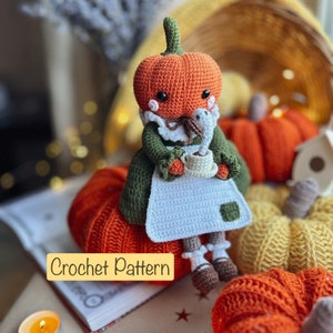 May include: Crochet pattern for a pumpkin-shaped amigurumi doll wearing a green dress and holding a cup. The doll is sitting on a book with other crocheted pumpkins in the background. The text "Crochet Pattern" is visible on the book.