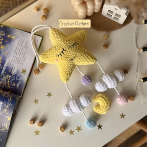 Könnte beinhalten: Ein handgefertigter gelber Häkelstern mit geschlossenen Augen und hängenden weißen Wolken, einem gelben Halbmond und pastellfarbenen Kugeln. Der Text "Crochet Pattern" ist sichtbar. Der Stern ist Teil eines Babymobils.