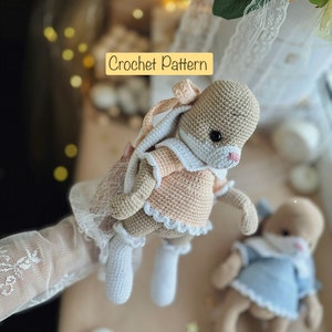 Può includere: Due bambole di coniglio all'uncinetto, una con un vestito color pesca e l'altra con un vestito blu. Entrambe le bambole hanno le mani alzate in aria. Il testo "Crochet Pattern" è visibile nell'immagine.
