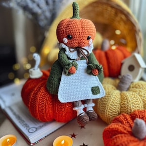 Op de afbeelding: Een gehaakte pompoendoll in een groene jurk en een witte schort zit op een grote oranje pompoen. Er zijn andere pompoenen op de achtergrond, waaronder een gele en een kleinere oranje.