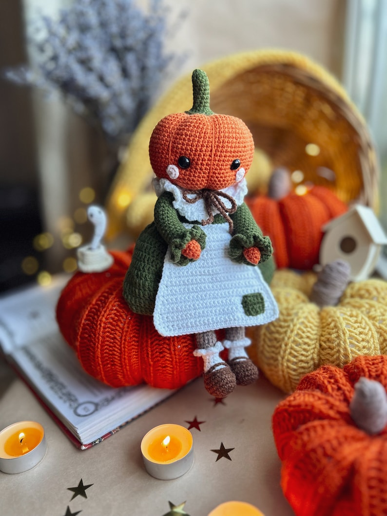Op de afbeelding: Een gehaakte pompoendoll in een groene jurk en een witte schort zit op een grote oranje pompoen. Er zijn andere pompoenen op de achtergrond, waaronder een gele en een kleinere oranje.