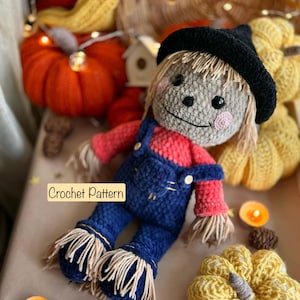 Può includere: Una bambola di spaventapasseri all'uncinetto con un cappello nero, una camicia rossa e una salopette blu. La bambola è circondata da zucche all'uncinetto e decorazioni autunnali.