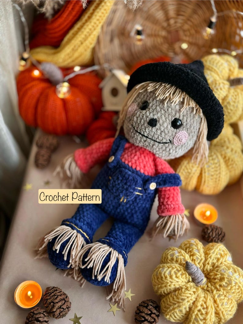 May include: A crocheted scarecrow doll wearing a black hat, red shirt, and blue overalls. The doll is surrounded by crocheted pumpkins and fall decor.