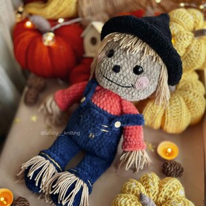 May include: A crocheted scarecrow doll wearing a black hat and blue overalls. The doll is surrounded by crocheted pumpkins and other fall decorations. The text "@lullaby__knitting" is visible in the image.