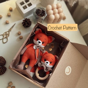 Puede incluir: Una caja de cartón marrón con un juguete de zorro de crochet y un anillo de dentición de madera en su interior. La caja está abierta y los juguetes son visibles. El texto "Crochet Pattern" está impreso en la caja. La palabra "Foxy" está escrita con fichas de letras sobre una superficie blanca.