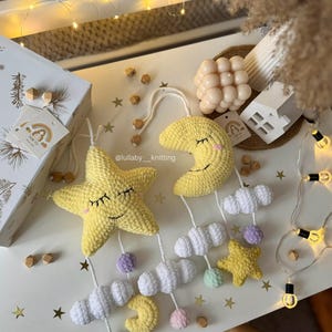 May include: A handmade crochet baby mobile featuring a yellow star and crescent moon with smiling faces, white clouds, and colorful pom-poms. The mobile is suspended from a white string and decorated with small wooden beads. The mobile is a nursery decoration.