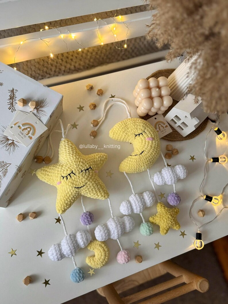 May include: A handmade crochet baby mobile featuring a yellow star and crescent moon with smiling faces, white clouds, and colorful pom-poms. The mobile is suspended from a white string and decorated with small wooden beads. The mobile is a nursery decoration.