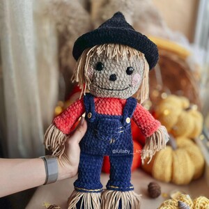 May include: A crocheted scarecrow doll wearing a black witch hat, red and orange shirt, blue overalls, and straw accents. The doll is being held by a hand with a silver watch.