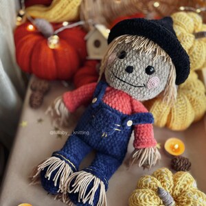 May include: A crocheted scarecrow doll wearing a black hat, red shirt, and blue overalls. The doll is surrounded by crocheted pumpkins and fall decor.