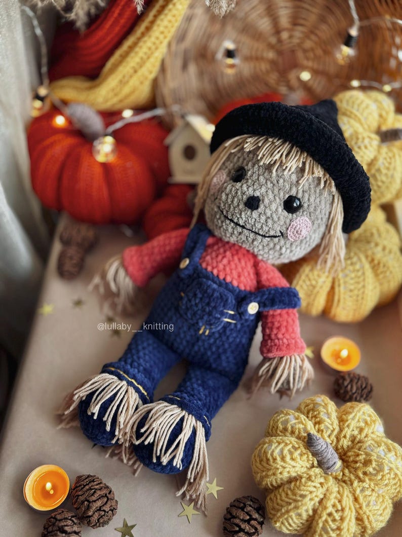 May include: A crocheted scarecrow doll wearing a black hat, red shirt, and blue overalls. The doll is surrounded by crocheted pumpkins and fall decor.