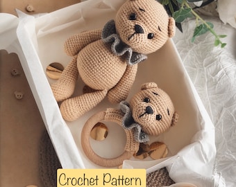 Könnte beinhalten: Zwei gehäkelte Teddybären, einer größer als der andere, sitzen in einer weißen Box mit einem Holzringspielzeug. Die Bären sind beige mit grauen Akzenten und schwarzen Augen. Die Box ist von einem weißen Band und goldenem Glitzer umgeben.