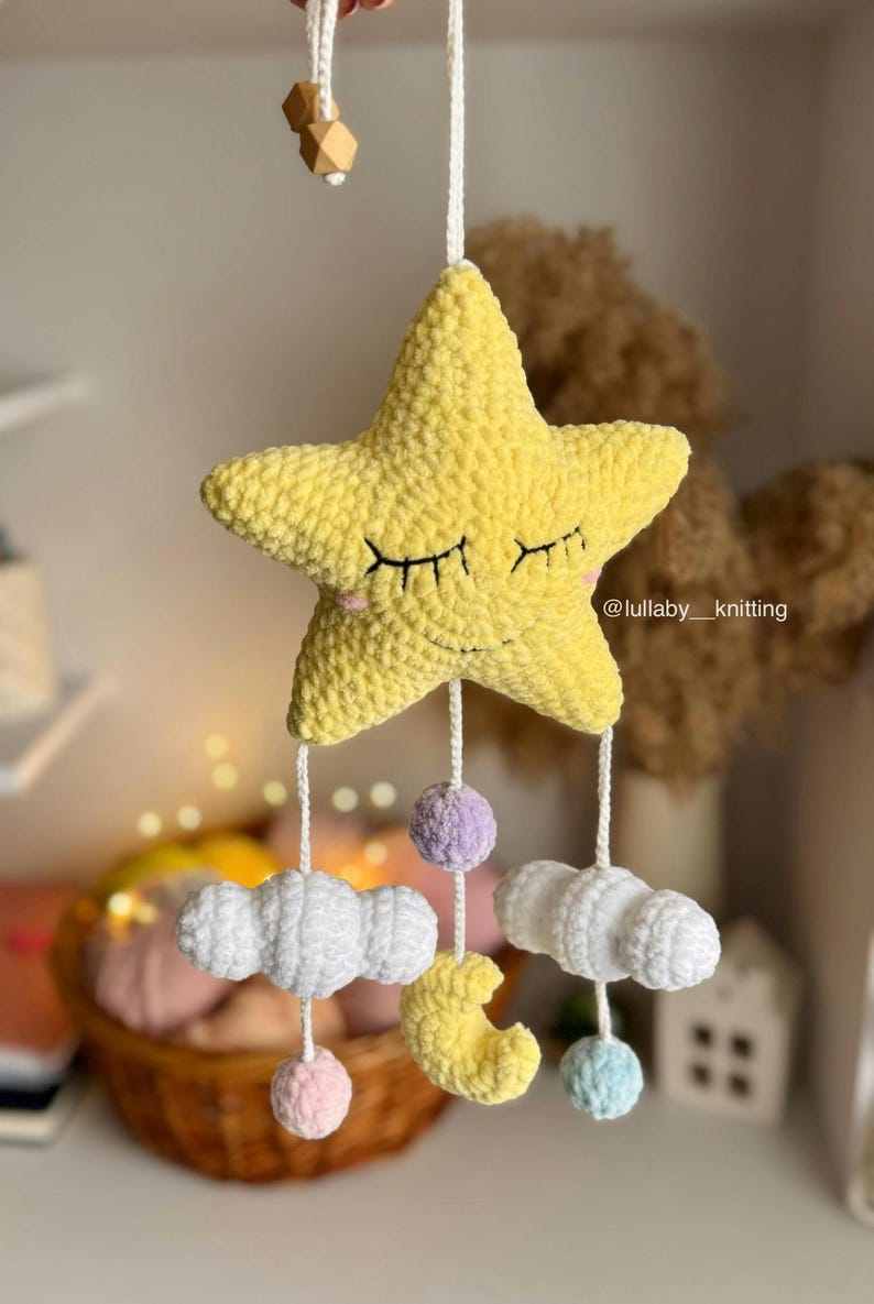 May include: A handmade yellow star-shaped baby mobile with closed eyes, hanging from a white string. Below the star are white clouds, a yellow crescent moon, and colorful pom-poms. The mobile is a nursery decoration.