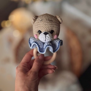 May include: A crocheted teddy bear rattle with a blue and white ruffle collar and a wooden ring. The rattle is held in a hand.