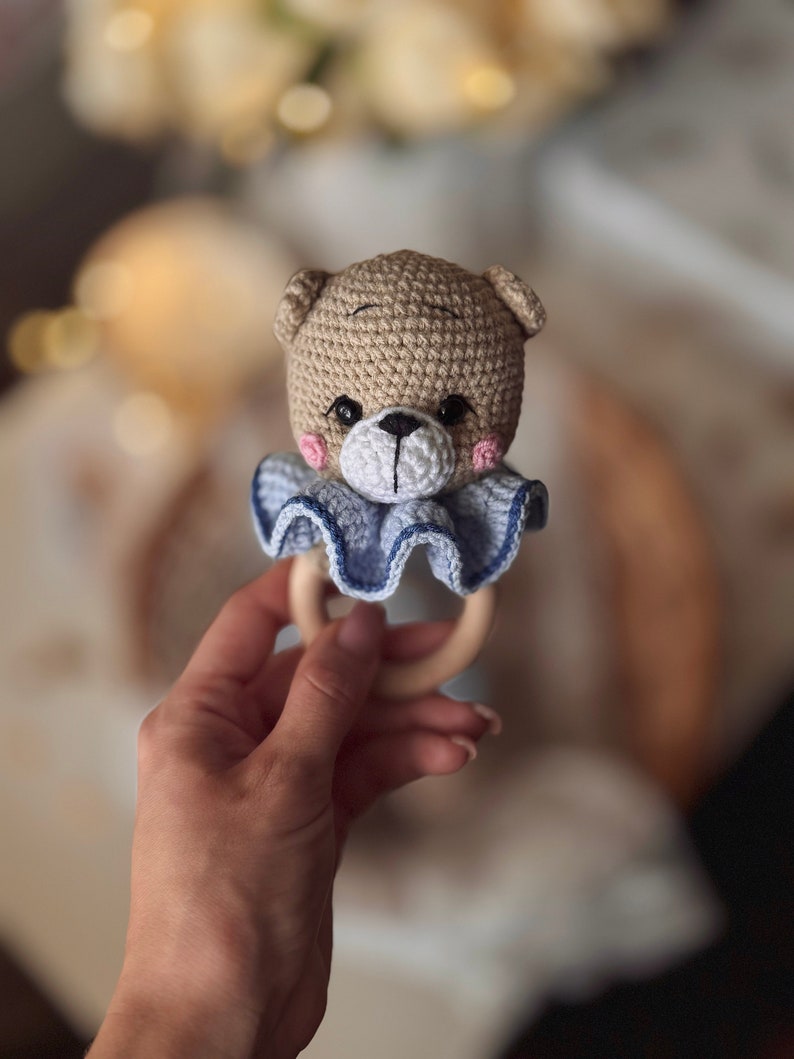 May include: A crocheted teddy bear rattle with a blue and white ruffle collar and a wooden ring. The rattle is held in a hand.