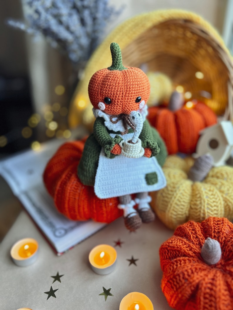 Op de afbeelding: Een gehaakte pompoendoll die een groene jurk draagt en een kopje vasthoudt. De pop zit op een grote oranje gehaakte pompoen. Er zijn andere gehaakte pompoenen op de achtergrond, evenals theelichtjes en sterren.