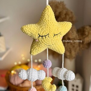 May include: A handmade yellow star-shaped baby mobile with closed eyes, hanging from a white string. Below the star are white clouds, a yellow crescent moon, and colorful pom-poms. The mobile is a nursery decoration.