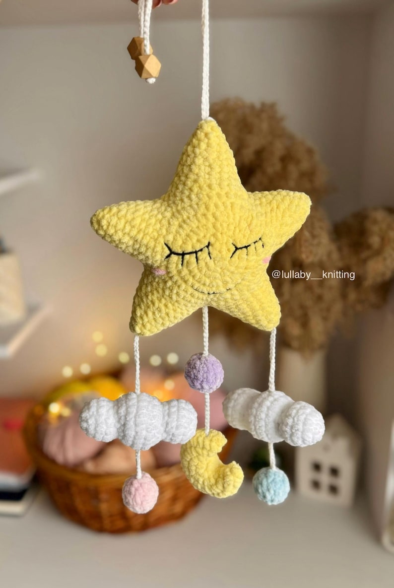 May include: A handmade yellow star-shaped baby mobile with closed eyes, hanging from a white string. Below the star are white clouds, a yellow crescent moon, and colorful pom-poms. The mobile is a nursery decoration.