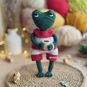 Pode incluir: Um sapo de crochê verde vestindo shorts listrados vermelho e branco segura uma caneca branca com líquido marrom. O sapo está em pé em um tapete de crochê marrom com uma pequena decoração de borboleta.