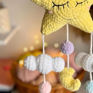 May include: A handmade baby mobile featuring a large yellow star with closed eyes, a smiling face, and hanging white clouds, a yellow crescent moon, and colorful pom-poms. The mobile is crafted from soft, textured yarn, creating a whimsical nursery decoration.