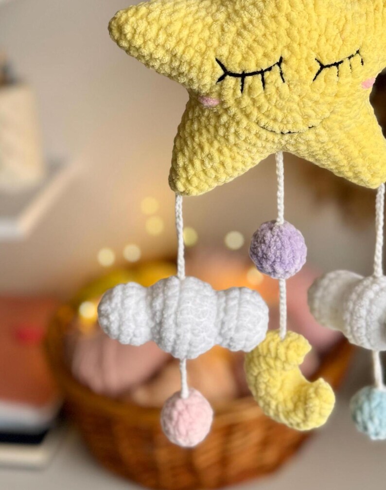 May include: A handmade baby mobile featuring a large yellow star with closed eyes, a smiling face, and hanging white clouds, a yellow crescent moon, and colorful pom-poms. The mobile is crafted from soft, textured yarn, creating a whimsical nursery decoration.