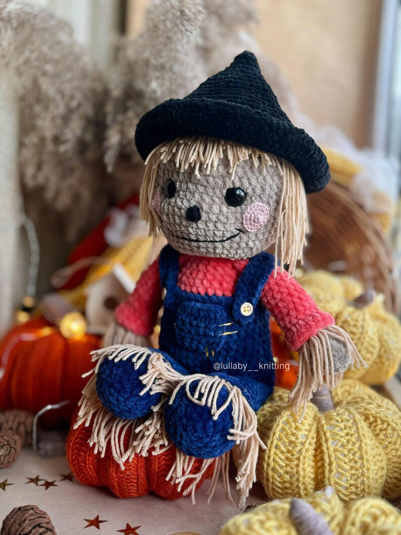 May include: A crocheted scarecrow doll wearing a black witch hat, red and blue overalls, and sitting on a crocheted pumpkin. The doll has a friendly face with brown eyes and pink cheeks. The doll is surrounded by crocheted pumpkins.