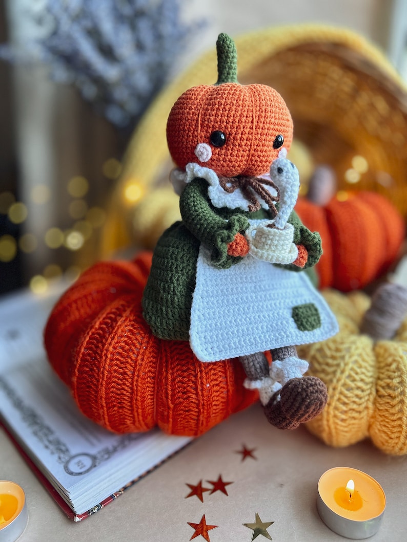 Op de afbeelding: Een gehaakte pompoendoll die een groene jurk en een witte schort draagt, een kopje vasthoudt en op een grote oranje gehaakte pompoen zit. Er zijn kleinere pompoenen op de achtergrond.