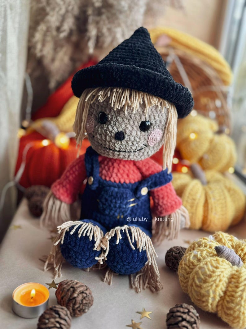 May include: A crocheted scarecrow doll wearing a black witch hat, red and blue overalls, and a straw-like beard. The doll is sitting on a table with pumpkins and pine cones.