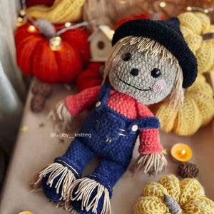 May include: A crocheted scarecrow doll wearing a black hat, red shirt, and blue overalls. The doll is surrounded by crocheted pumpkins and fall decor.